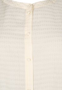 Blusa color crema con texture a griglia, dotata di chiusura a bottoni sul davanti e realizzata in un tessuto morbido e leggero.