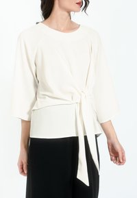 Top blanco con mangas tres cuartos y detalle de lazo en la cintura. Tejido suave y liso con un corte relajado, combinado con pantalones negros.