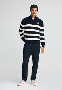 Stickad tröja i marinblå och beige ränder, med hög krage och halvdragkedja. Bärs med mörka byxor och ljusa sneakers.