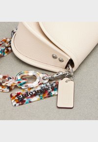 Torba ze skóry o teksturze w odcieniu jasnobrązowym z zaokrąglonym kształtem, wyposażona w wielokolorowy łańcuch akrylowy oraz srebrne okucia, w tym zawieszkę z logo.