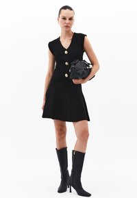 Schwarzes ärmelloses Kleid mit enganliegendem Oberteil und ausgestellt Rock, mit goldenen Knöpfen. Eine schwarze Handtasche mit goldenen Akzenten wird gehalten. Knielange schwarze Stiefel.