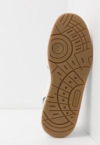 Semelle de basket Lacoste, en caoutchouc marron texturé avec un motif circulaire et linéaire, mettant en avant durabilité et adhérence. Partie supérieure blanche visible.