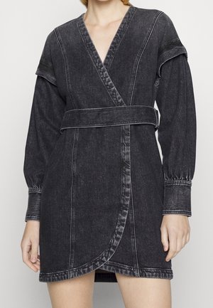 Kobieta ubrana w wyblakłą czarną sukienkę jeansową z długimi bufkami i pasem w talii.