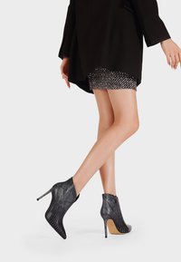 Stivaletti alla caviglia in pelle di serpente nera con alti tacchi a stiletto, dotati di chiusura con zip e motivo testurizzato. Abbinati a una mini gonna scintillante argento.