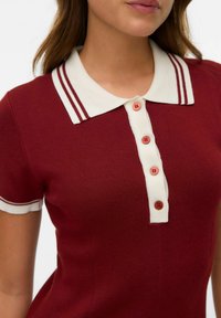 Röd ribbad polo med krämfärgad krage, försedd med två röda ränder och fem knappar med färgade knappar.