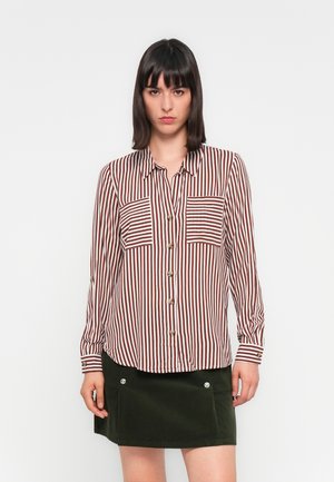 Woman with straight dark hair wearing a brown and white vertical striped button-up shirt and a dark green skirt with front pockets.
