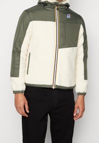 Gepolsterte Jacke mit olivgrünem Oberteil und cremefarbenem Fleece-Unterteil. Verfügt über einen Reißverschluss vorne und einen bunten gestreiften Reißverschluss sowie elastische Bündchen.