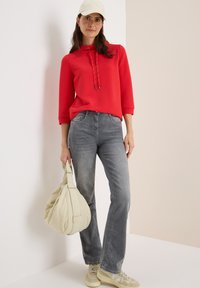 Rood top met structuur, voorzien van een capuchon en trekkoorden, gecombineerd met grijze flared jeans. Draagt een crèmekleurige tas en beige sneakers.