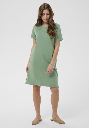 Žena s dlhými vlnitými vlasmi, oblečená v krátkom zelenom šate s rukávmi po lakte a béžových pletených nazúvacích topánkach, stojaca s mierne sklonenou hlavou.