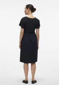 Schwarzes knielanges Kleid mit kurzen Puffärmeln, elastischem Bund und weicher Textur. Gestylt mit schwarzen flachen Schuhen und minimalem Zubehör.