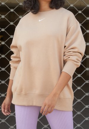 Person iført en oversized beige Nike-sweatshirt og ribbede lavendelfarvede leggings, stående foran en metalkædeport.