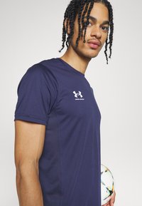 Námornícky modré športové tričko s krátkymi rukávmi, vyrobené z hladkej tkaniny. Na hrudi je biele logo Under Armour. Futbalová lopta je držená na boku.