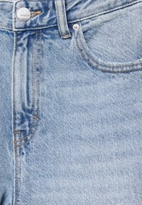 Jean en denim bleu clair en gros plan montrant la poche avant, la boucle de ceinture, le bouton en métal et les détails de couture.