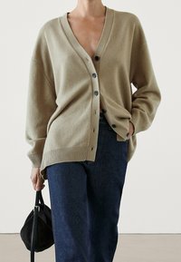 Beige oversized cardigan with a V-neck and five dark buttons, ribbed hem, worn over dark blue jeans, held with a black small bag.