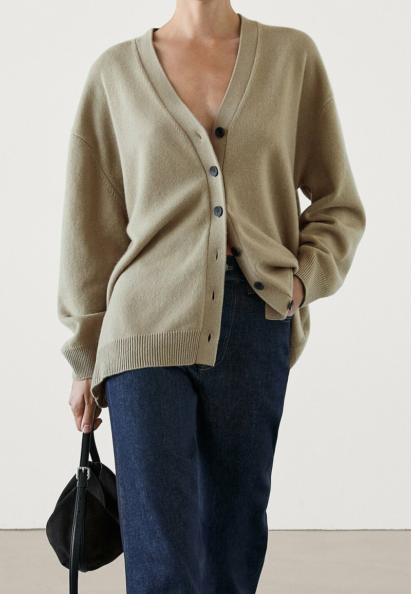 Beige oversized cardigan with a V-neck and five dark buttons, ribbed hem, worn over dark blue jeans, held with a black small bag.