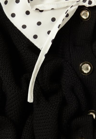 Tejido negro de punto con textura visible, superpuesto con una tela de satén blanca con puntos. Presenta botones dorados y un cuello detallado.