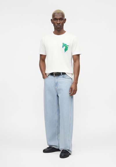 Άνδρας με κοντά ξανθά μαλλιά που φοράει λευκό μπλουζάκι με πράσινο-μπλε σχέδιο ψαριού, ανοιχτό μπλε φαρδιά τζιν παντελόνια, μαύρη ζώνη και μαύρα παπούτσια.