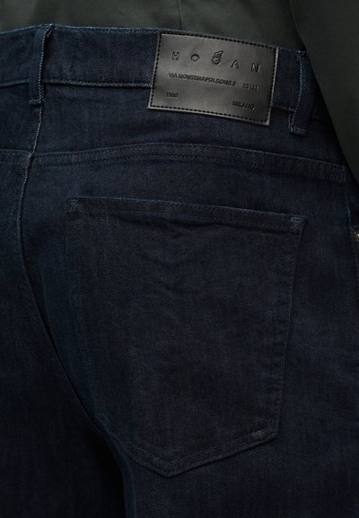 Πίσω τσέπη σκούρου μπλε τζιν με ραμμένη ετικέτα που γράφει «Hogan» και λεπτομέρειες διεύθυνσης σε μαύρο στο στρίφωμα της μέσης.