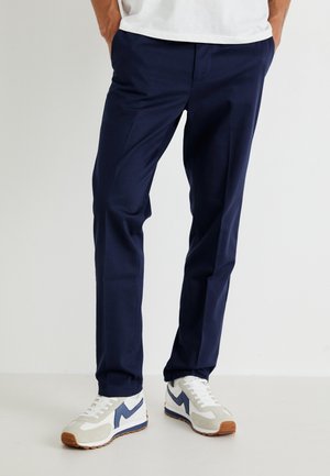 Man wearing navy blue tailored pants and white sneakers with blue and brown accents, standing with hands in pockets.