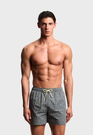 Uomo muscoloso con capelli corti e scuri che indossa pantaloncini da bagno grigi con lettere bianche e un laccio giallo, in piedi contro uno sfondo semplice.