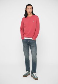 Sweatshirt i korall, rund halsringning, ribbade ärmslut, tillsammans med ljusblå denimjeans och gröna sneakers med vita detaljer.