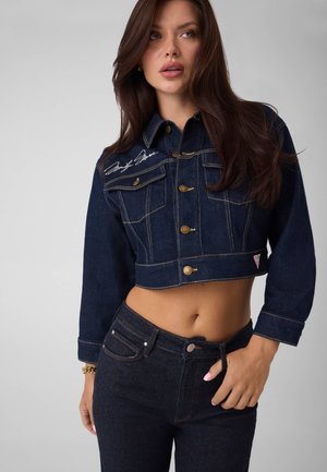 Femme aux longs cheveux foncés porte une veste courte en jean foncé avec des boutons dorés et un jean taille haute en jean foncé assorti.