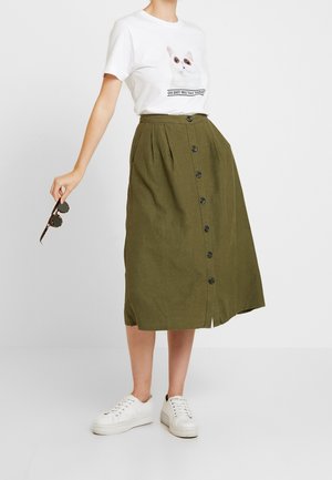 Nainen, joka pukeutunut valkoiseen t-paitaan, jossa on kissa-aiheinen grafiikka, oliivinvihreään napitettavaan midihameeseen ja valkoisiin lenkkareihin, pitäen aurinkolaseja oikeassa kädessään.