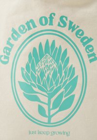 Borsa in tela beige con testo turchese e un'illustrazione botanica, con la frase "Giardino di Svezia" sopra e "continua a crescere" sotto.