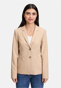 Beżowa marynarka z gładkiego materiału, z klapami, z dwoma guzikami z przodu i dwiema kieszeniami. Noszona na białej bluzce.