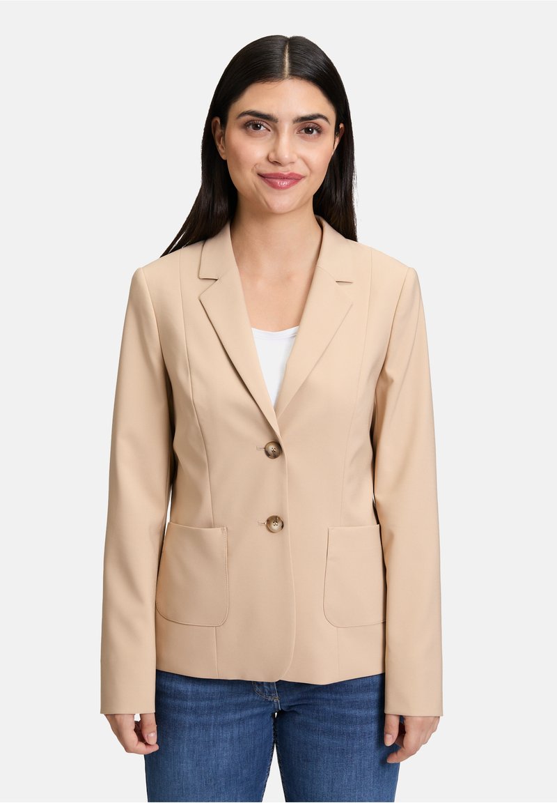 Beżowa marynarka z gładkiego materiału, z klapami, z dwoma guzikami z przodu i dwiema kieszeniami. Noszona na białej bluzce.
