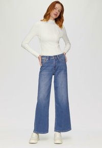 Witte lange mouwtop met ribbelstructuur, gecombineerd met wijde blauwe jeans. Model draagt lichtgekleurde sneakers. Simpel, schoon ontwerp.