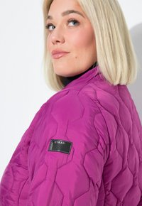 Žena s blond bob účesem, oblečená v fialové prošívané bundě, se dívá přes rameno na bílém pozadí.