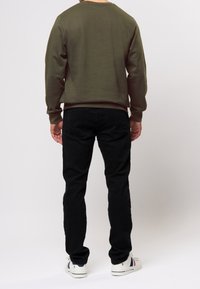 Oliivinvihreä huppari pyöreällä kaula-aukoella, yhdistettynä mustiin housuihin. Kangas näyttää pehmeältä ja tasaiselta, ja asua korostaa valkoiset lenkkarit.