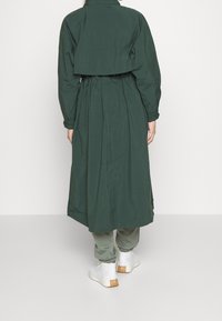 Zelený oversized trenčkot se stáhnutým pasem, dlouhými rukávy a bočními rozparky, v kombinaci se světle zelenými kalhotami a bílými botami.