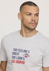 T-shirt en coton gris avec des lettres audacieuses en rouge et bleu, affichant le texte "LE SENTIMENT EST SUPER, LE LOOK EST LEE COOPER."