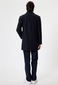 Marineblauw wollen blend jas met een rechte snit, brede kraag, twee zijzakken en een centrale rugsplit; gecombineerd met donkere broeken en witte sneakers.