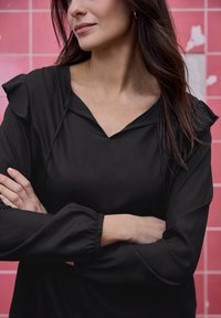 Blouse noire avec décolleté en V, épaules à volants et manches froncées. Texture lisse sur un fond contrastant de carreaux roses.