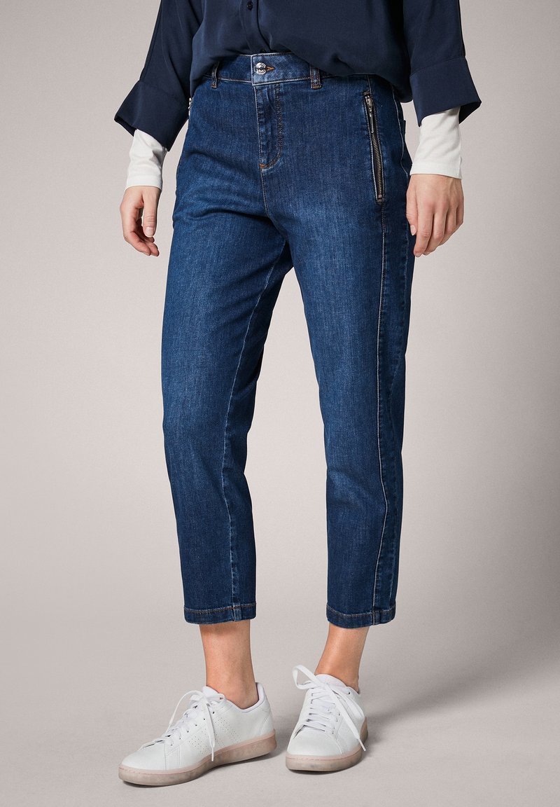Femme portant un pantalon court en denim bleu avec des poches zippées sur les côtés, un haut bleu marine avec des manches blanches, et des baskets blanches à lacets.