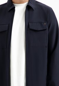 Marineblauwe jas met een gladde textuur, twee borstzakken en een puntige kraag. Draagt over een wit shirt, met een ritssluiting.