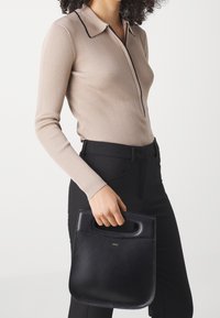 Kvinde i en beige ribbet langærmet top og sorte bukser, der holder en sort lædertaske med håndtag i udsnit mod en hvid baggrund.