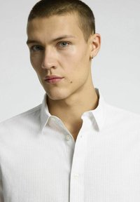 Vit skjorta med knapp-up i en subtil texturerad mönster, med krage och vita knappar, uppvisad på en modell med en neutral bakgrund.