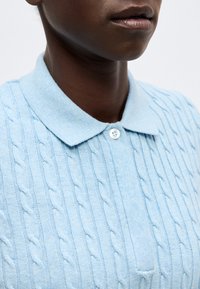 Άτομο με σκουρόχρωμο δέρμα που φορά ένα γαλάζιο πουλόβερ πλεκτό πόλο με ένα λευκό κουμπί κλεισμένο στον γιακά.