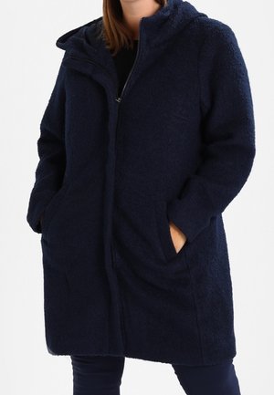 Personne portant un manteau zippé texturé bleu foncé avec une capuche et les mains dans les poches avant, visible des épaules jusqu'au milieu des cuisses.