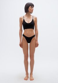 Γυναίκα με κοντά σκούρα μαλλιά, όρθια ξυπόλυτη, φορώντας μαύρο μπραλέτο και αντίστοιχο ψηλοκόφτη εσώρουχο, κοιτάζει προς τα δεξιά σε απλό λευκό φόντο.