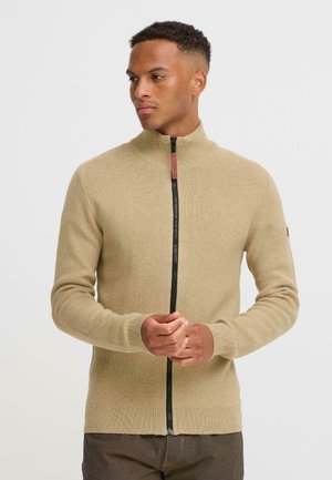 Suéter de punto beige con un alto cuello y una cremallera negra de longitud completa. Presenta puños y dobladillo de canalé, diseñado con una textura suave.