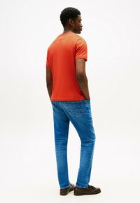 Camisa de tirantes corta para hombre en color naranja, de corte casual, combinada con jeans de mezclilla azul y zapatos oscuros. Diseño simple con una textura suave y líneas limpias.