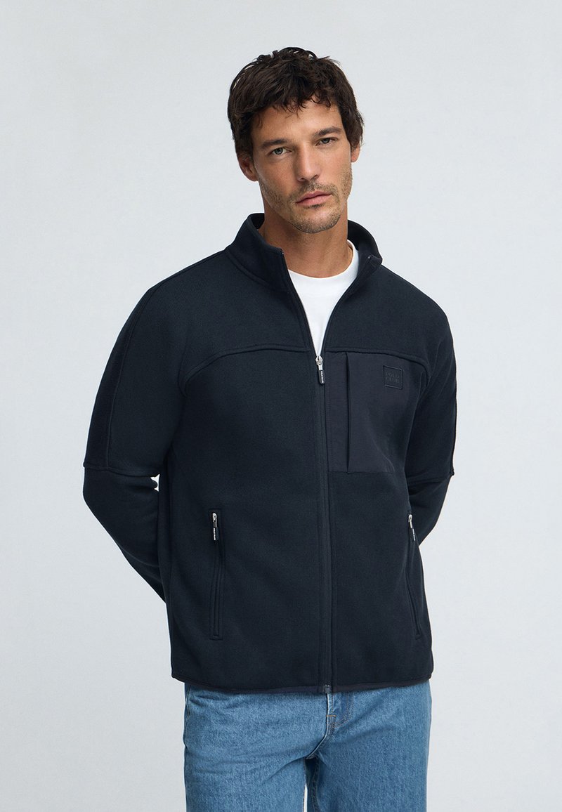 Svart fleecejacka med hög krage, framdragkedja och bröstficka. Har två sidofickor och en mjuk textur.