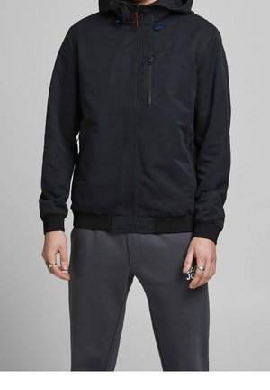 Homme portant une veste noire à capuche zippée avec une poche poitrine zippée et un pantalon de survêtement gris foncé avec un logo blanc sur la cuisse gauche.