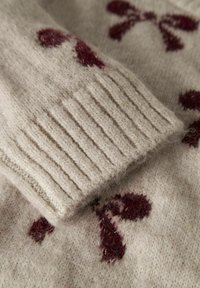 Détail d'un pull en tricot beige avec des poignets côtelés, orné d'un nœud bordeaux et de motifs à pois. Matériau doux et texturé avec un léger duvet.