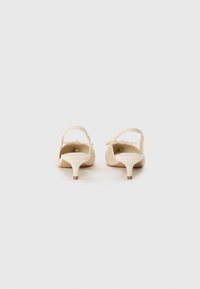Beige heeled sandals with an open back, featuring a thin heel, bow accent, and smooth texture. The sole has a subtle brown detail.